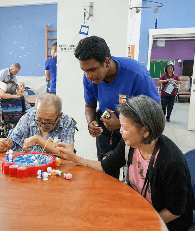 A caregiver in blue assists two elderly people playing a toy fishing board game at a table in a rehab area.